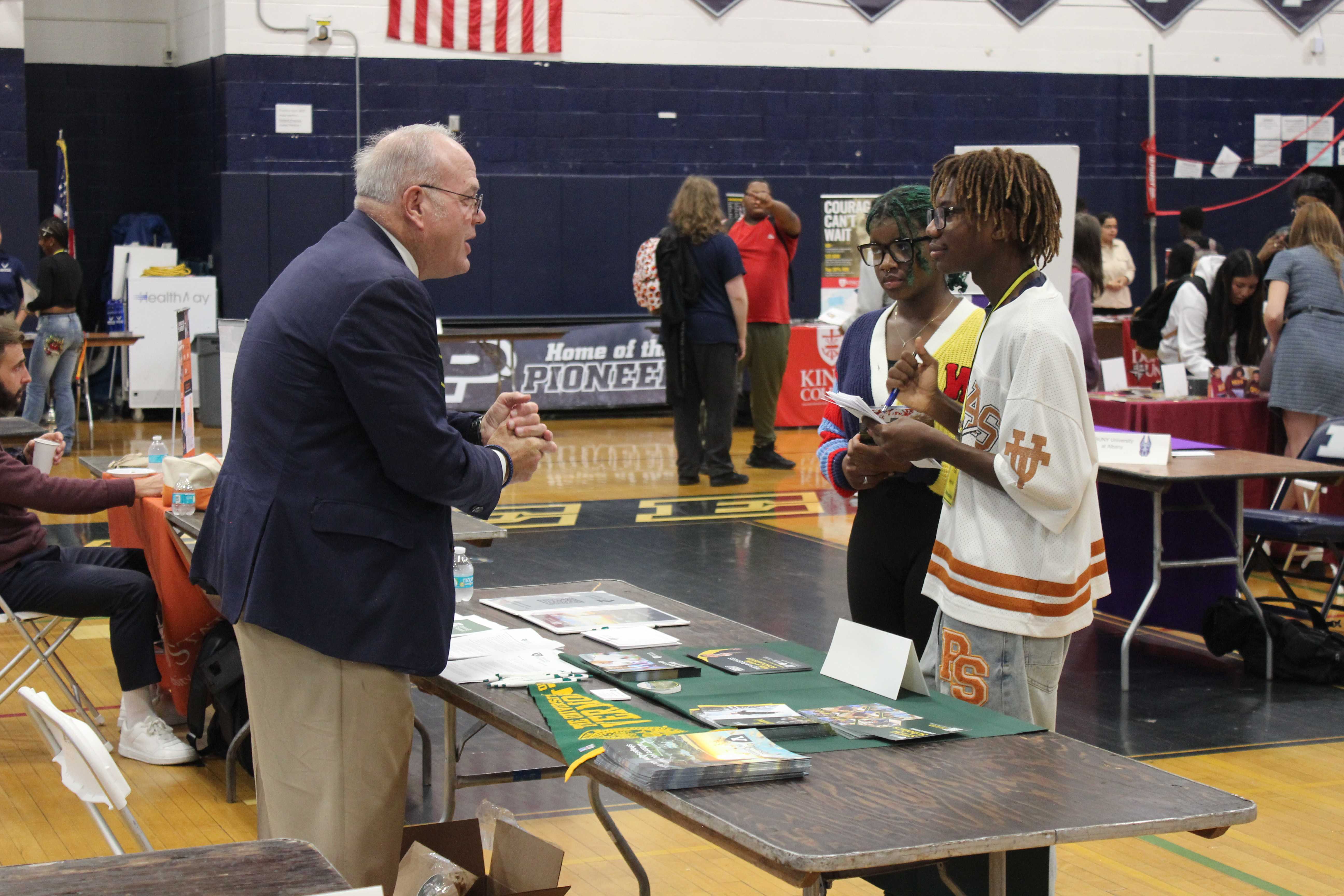 Two students speak to a man in a suit at a table at a college fair in a gymnasium